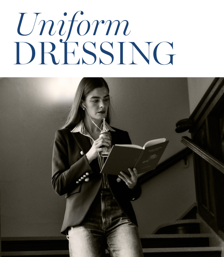 Black and white image of a woman walking down a flight of stairs reading a book and drinking a soda. Woman is in a blazer and jeans. Copy says: Uniform dressing. 