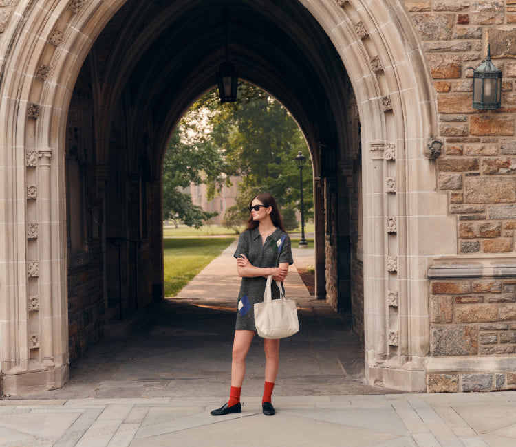 Woman standing in front of a stone arch in a short sleeve argyle sweater dress holding a cream tote bag. 