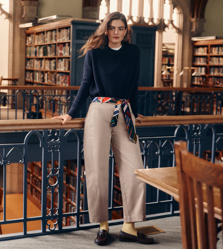 Woman standing in a library in a navy sweater, khakis, and a tie belt. 