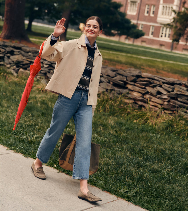 Woman walking on a campus with an umbrella in a cropped khaki tench coat, striped sweater, and jeans. 