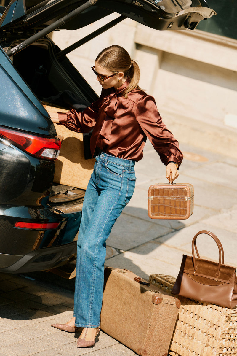 Woman in tortoiseshell sunglasses effortlessly holds a small travel case, wearing classic 90's Straight high-rise jeans, epitomizing fun, high-end style beside a dark car.
