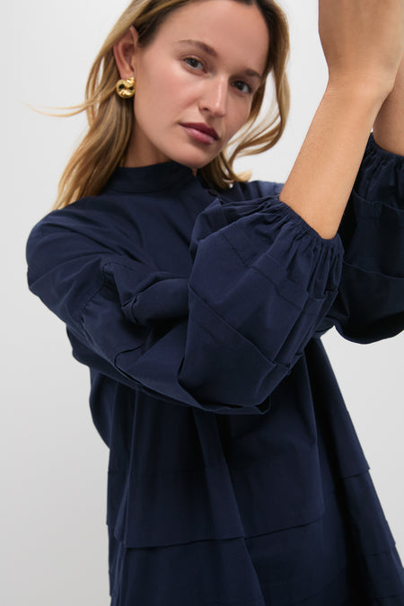 A woman models a Pomander Place classic navy long-sleeve top with tiered panel detailing, gold hoop earrings, and one arm raised against a plain light background.