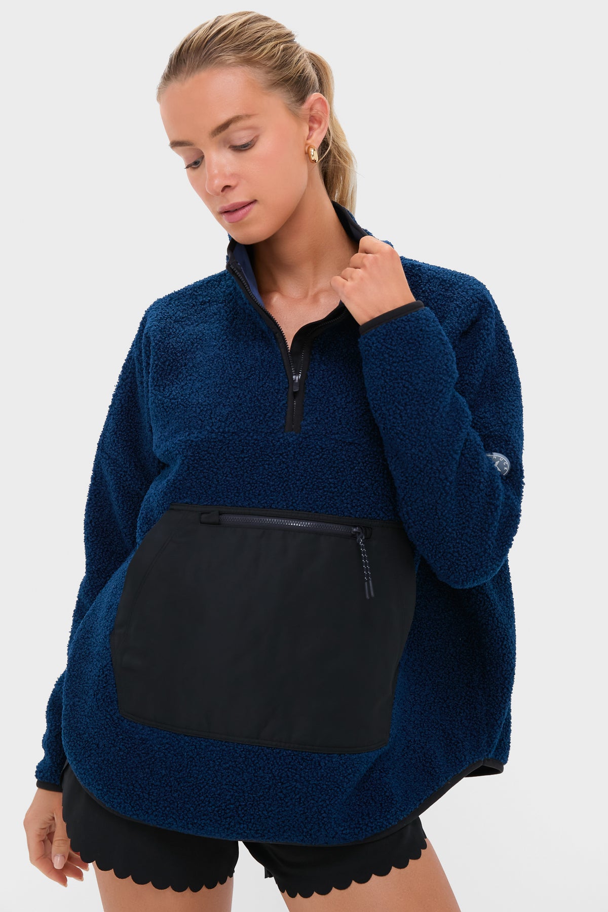 A woman wears a Tnuck Sport blue fleece pullover with a quarter-zip collar and large black front pocket, paired with black scalloped shorts, standing against a plain backdrop.