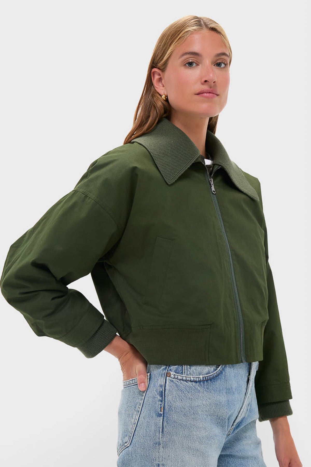 A person wearing a Sea New York green bomber jacket with a wide collar and light blue jeans stands against a plain light background.