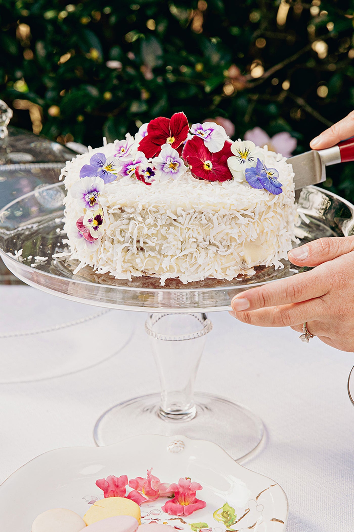Isabella Cake Stand: Classic glass pedestal elegantly displaying a coconut-frosted cake with edible flowers, embodying effortless, fun, high-end dining.