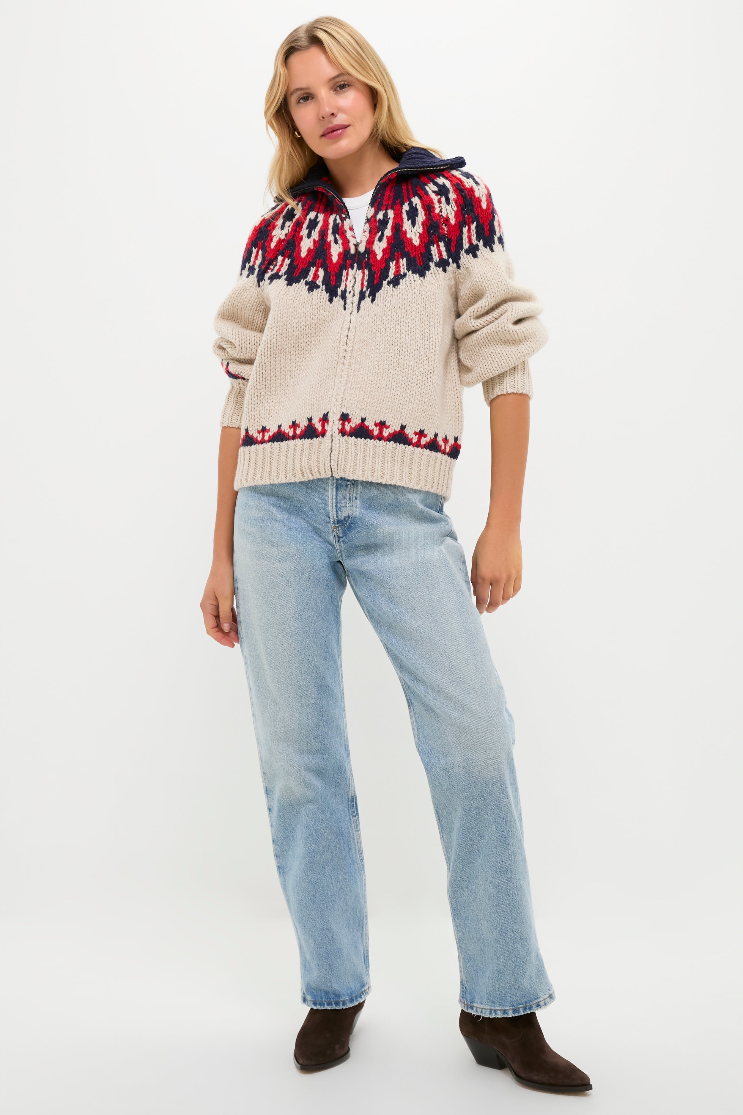 A person wears THE GREAT. Outdoors beige zip-up wool sweater with a red and navy patterned yoke, paired with light blue jeans and dark brown boots, standing against a plain white background.