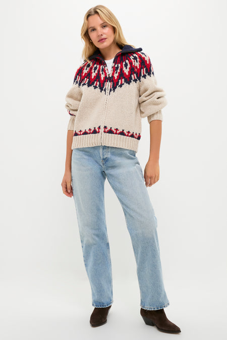 A person wears THE GREAT. Outdoors beige zip-up wool sweater with a red and navy patterned yoke, paired with light blue jeans and dark brown boots, standing against a plain white background.