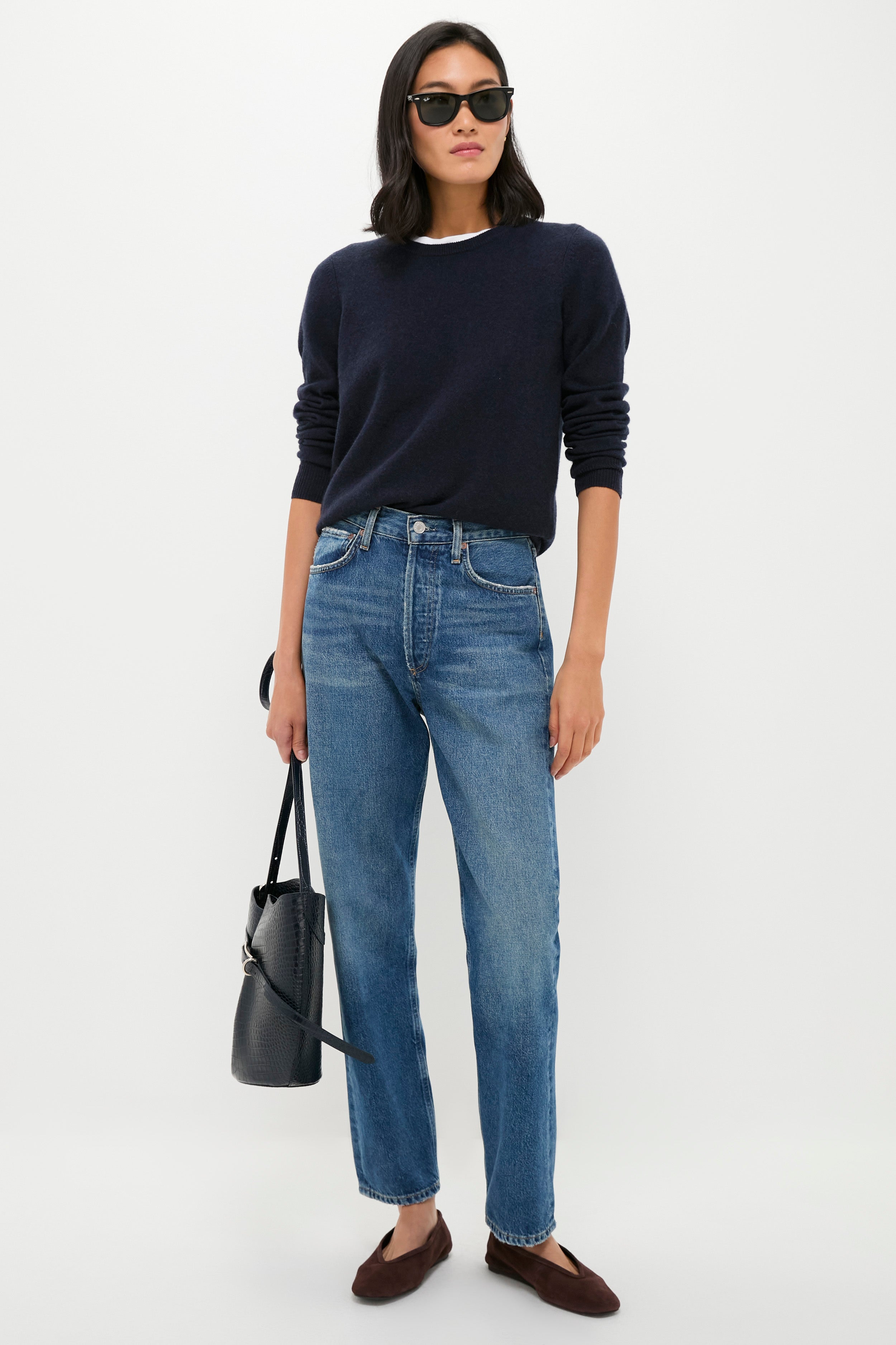 A woman wearing sunglasses, a Sandstone Cashmere Sweater from Tuckernuck, blue jeans, and brown flats stands holding a black textured handbag against a plain white background.