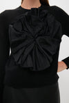 A woman wears a luxury black crewneck sweater featuring a large textured satin flower on the front, styled with a black skirt.