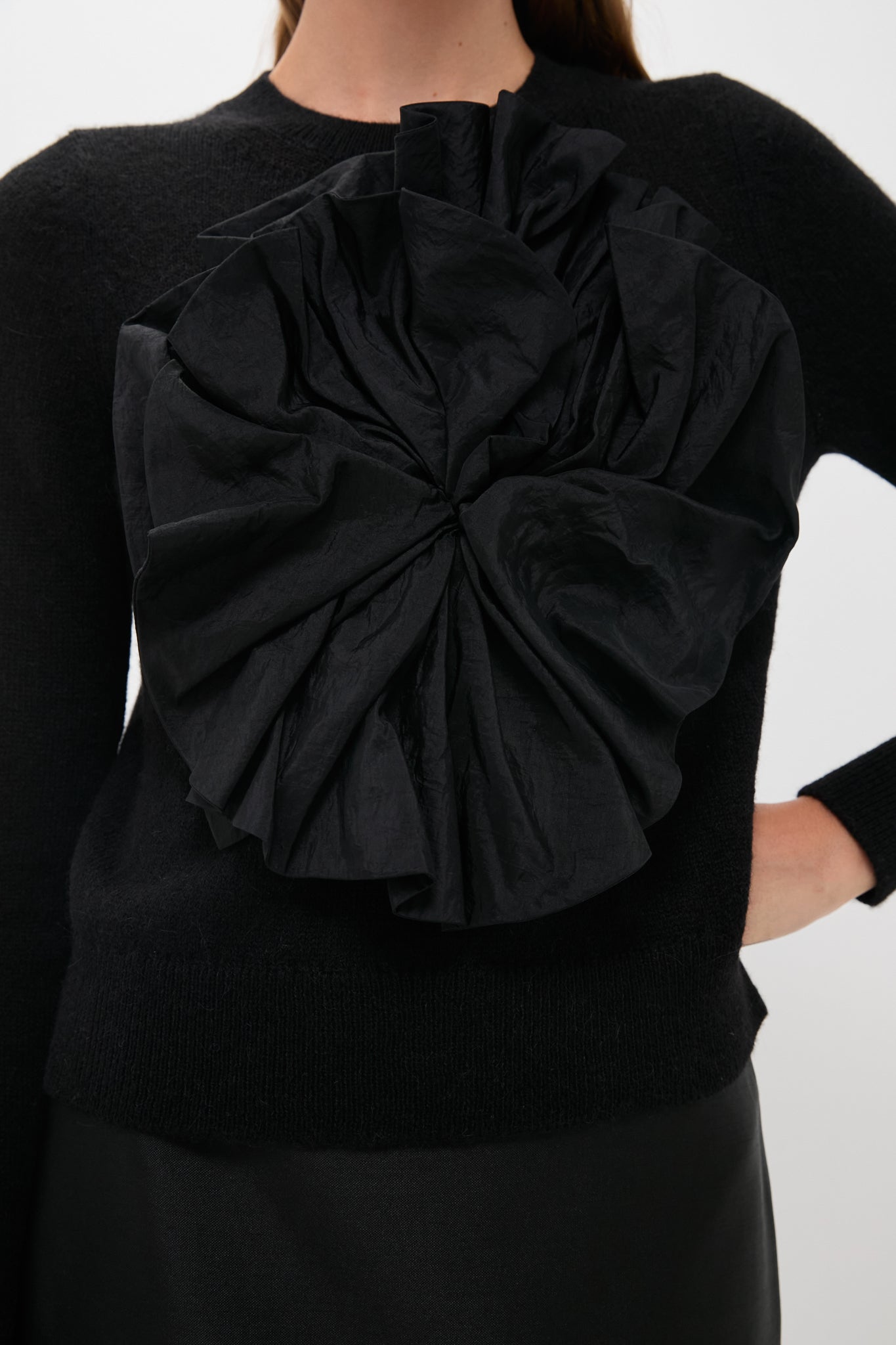 A woman wears a luxury black crewneck sweater featuring a large textured satin flower on the front, styled with a black skirt.