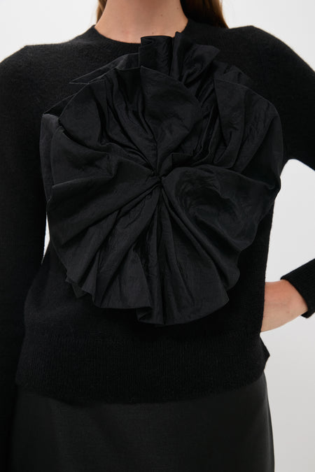 A woman wears a luxury black crewneck sweater featuring a large textured satin flower on the front, styled with a black skirt.