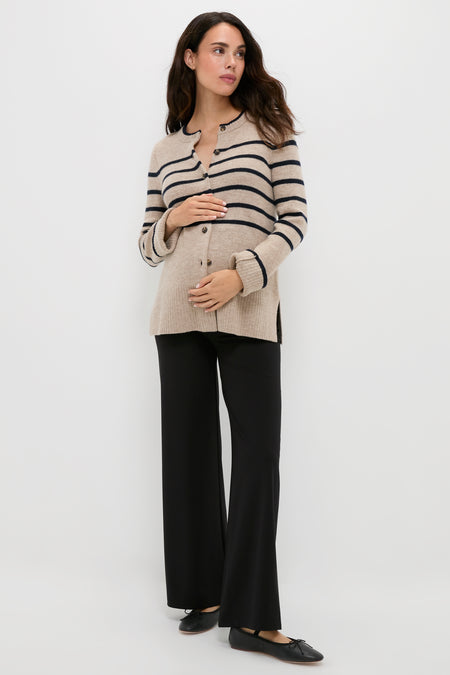 A woman stands against a plain white background, wearing a HATCH beige and black striped maternity sweater, black wide-leg pants, and black shoes, with one hand on her stomach.
