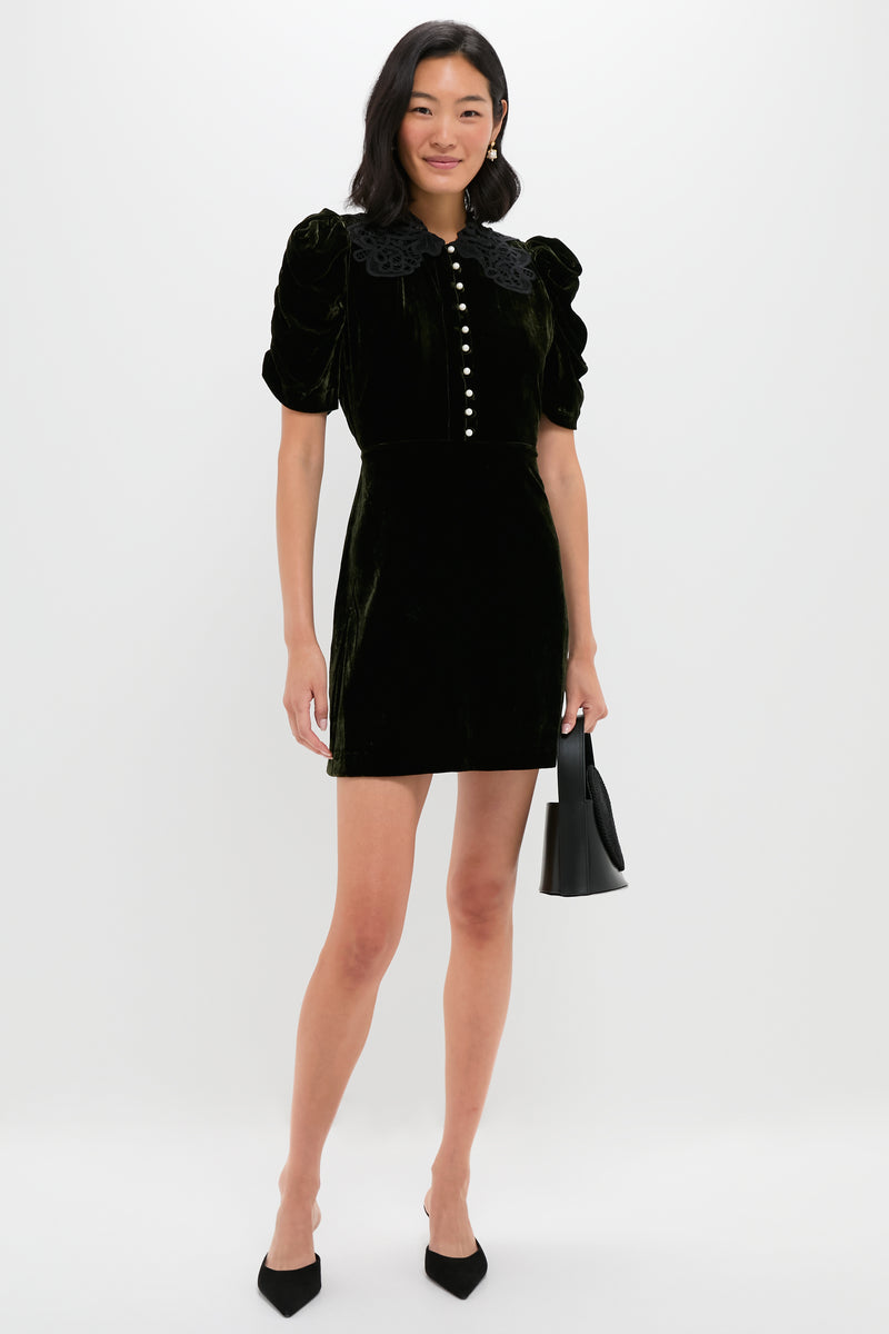 A woman poses against a white background in a Sea New York velvet mini dress featuring puff sleeves, a lace collar, and front buttons, styled with a small black handbag and black heels.