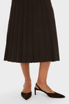 A person wearing a black STAUD midi skirt with pleats and black pointed-toe slingback heels stands against a white background.
