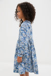 A young girl with curly hair stands sideways in a blue Rachel Riley floral print button-front dress against a plain white background.