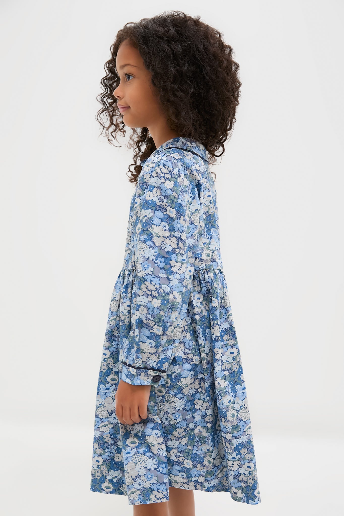 A young girl with curly hair stands sideways in a blue Rachel Riley floral print button-front dress against a plain white background.