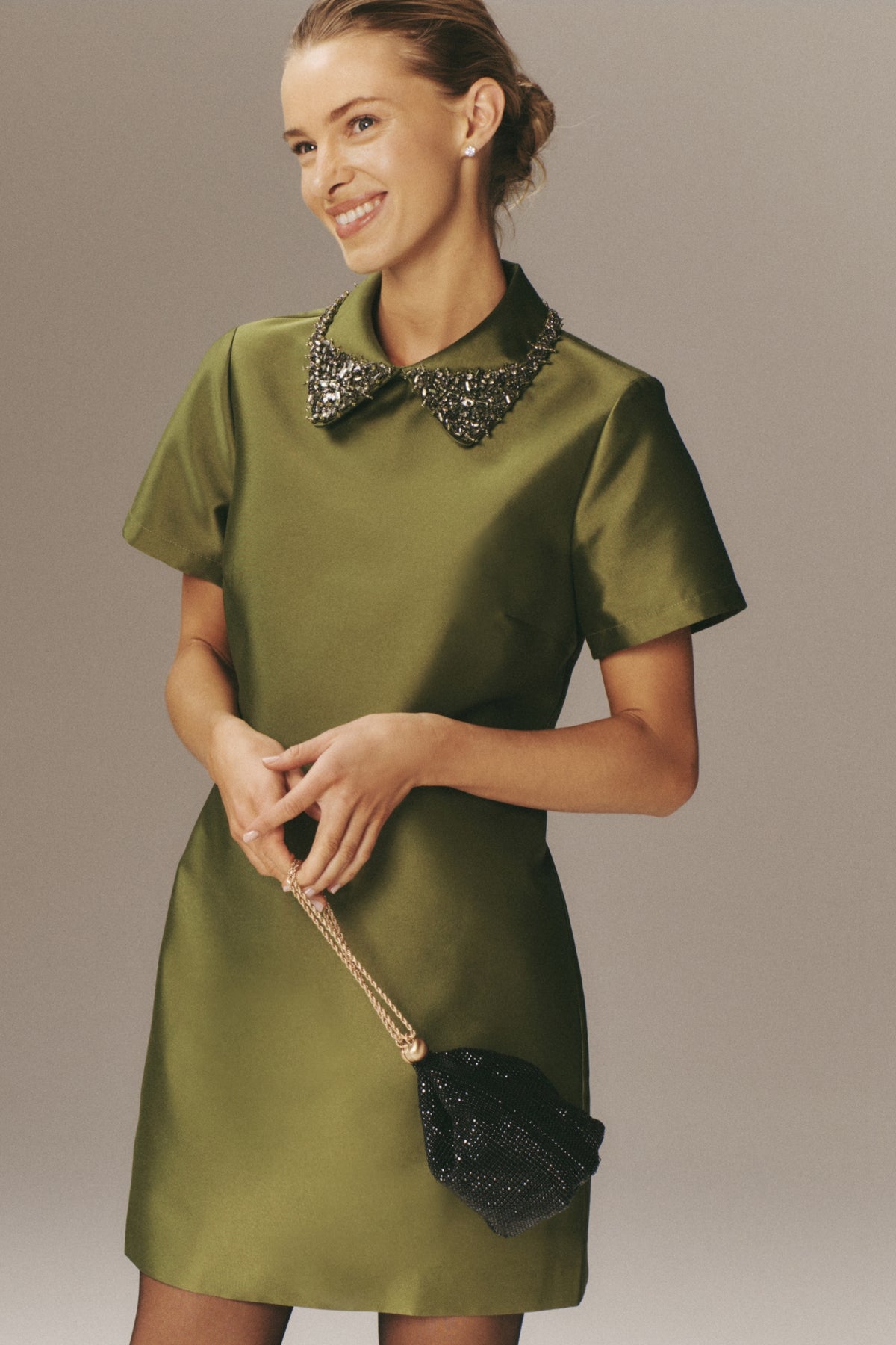 Smiling woman in a classic, effortless Green Embellished Aurelie Shift Dress with jeweled collar, holding a sparkly black clutch. Perfect for a fun, high-end party look.