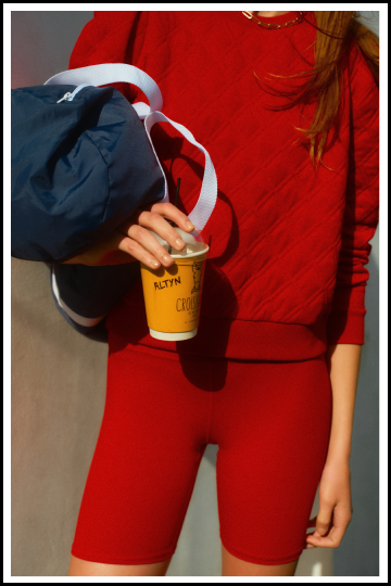 Closeup of a woman holding a coffee and a navy sports bag in a red sweatshirt and red bike shorts. 