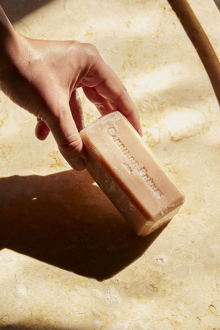 Hand picking up a lathered Florae Solid Body Soap, embossed with letters, over a stone sink. The vegan, floral-scented soap offers a classic, high-end bathing experience.