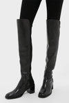A person in black stretch leather, low block heel over-the-knee boots and black leggings stands against a plain background, showcasing the signature Stuart Weitzman style.