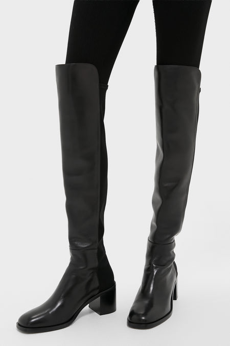A person in black stretch leather, low block heel over-the-knee boots and black leggings stands against a plain background, showcasing the signature Stuart Weitzman style.