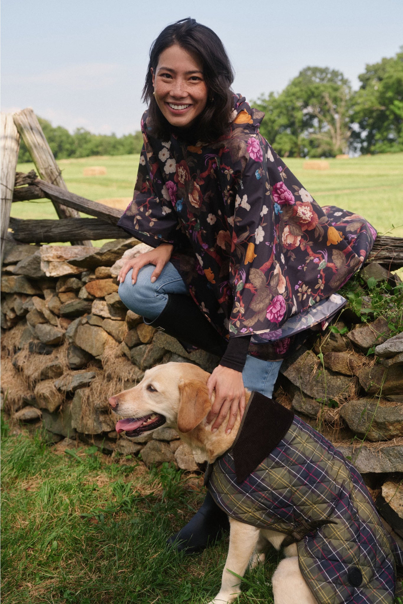 Woman sitting on a fence petting a dog. The woman wears a floral poncho. The dog wears a plaid jacket. 