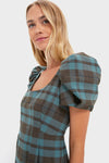 A woman wearing a Tuckernuck green and blue plaid column dress with puff sleeves stands against a plain light background.