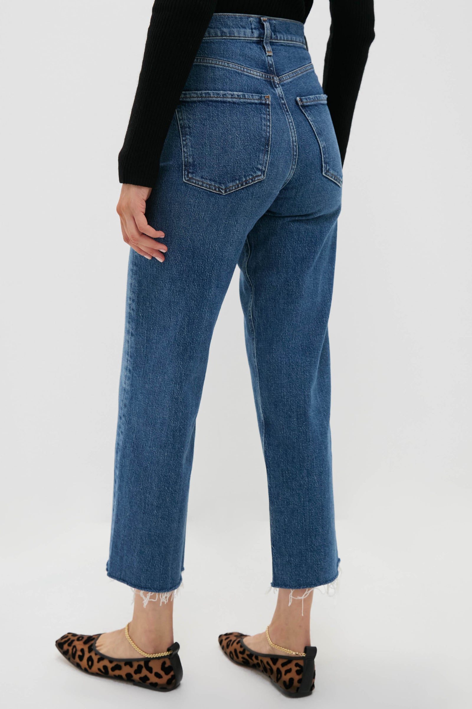 A person is shown from the back wearing AGOLDE high-rise straight-leg blue jeans with frayed hems, a black long-sleeve top, and leopard-print flats.