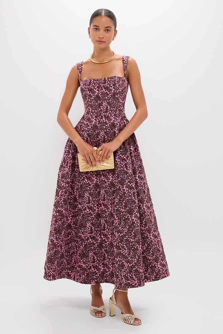 A woman wears the CLEA Sylvie Jacquard Dress—sleeveless, ankle-length, pink and black—styled with a gold clutch, gold sandals, and a gold necklace.