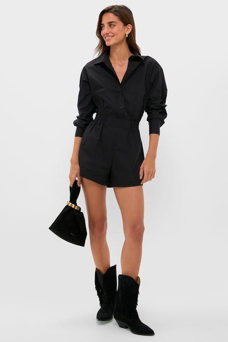 A woman wears the Hyacinth House Hartwell Playsuit— a long-sleeve black romper with a collared neckline—styled with black suede ankle boots and a small black handbag.