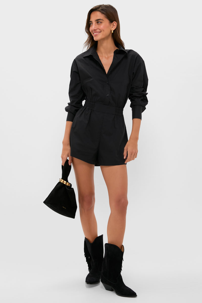 A woman wears the Hyacinth House Hartwell Playsuit— a long-sleeve black romper with a collared neckline—styled with black suede ankle boots and a small black handbag.