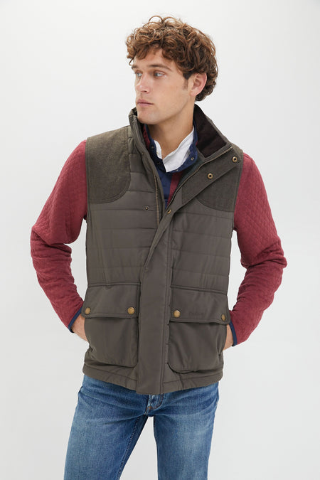 A man stands with his hands in his pockets, looking to the side. He wears a red quilted jacket layered with a brown Barbour gilet and blue jeans.