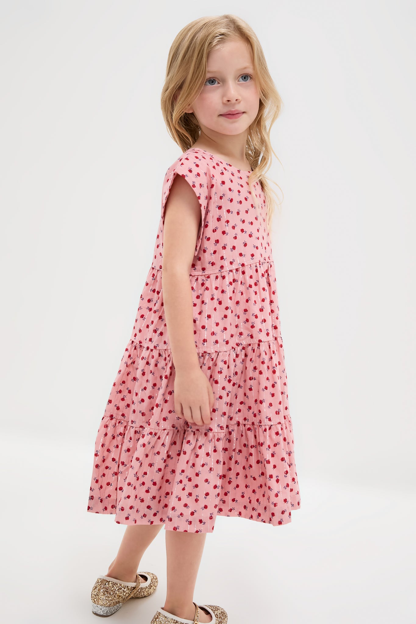 Young girl models the classic Girls Peachy Dress, featuring an effortless tiered silhouette and fun red floral print, paired with high-end sparkly Mary-Jane shoes.
