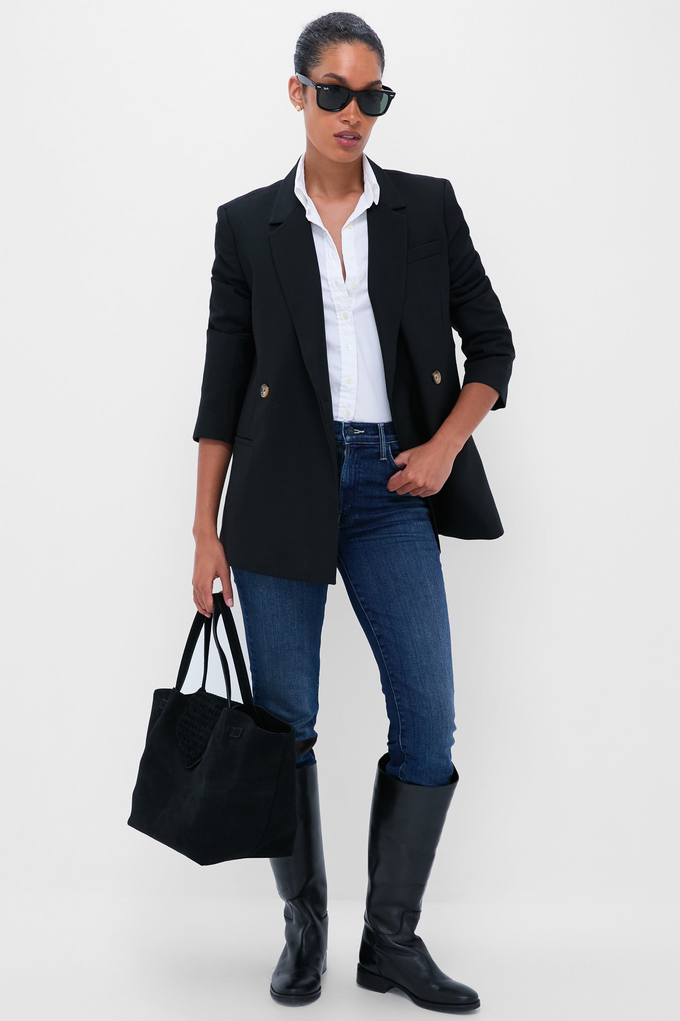 Woman in a black blazer, white shirt, MOTHER mid-rise straight leg jeans, knee-high black boots, sunglasses, and holding a black tote bag stands against a plain white background.