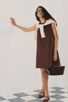 Woman in a brown suede shift dress, a cream sweater over her shoulders, and holding a chocolate brown mini tote bag. 