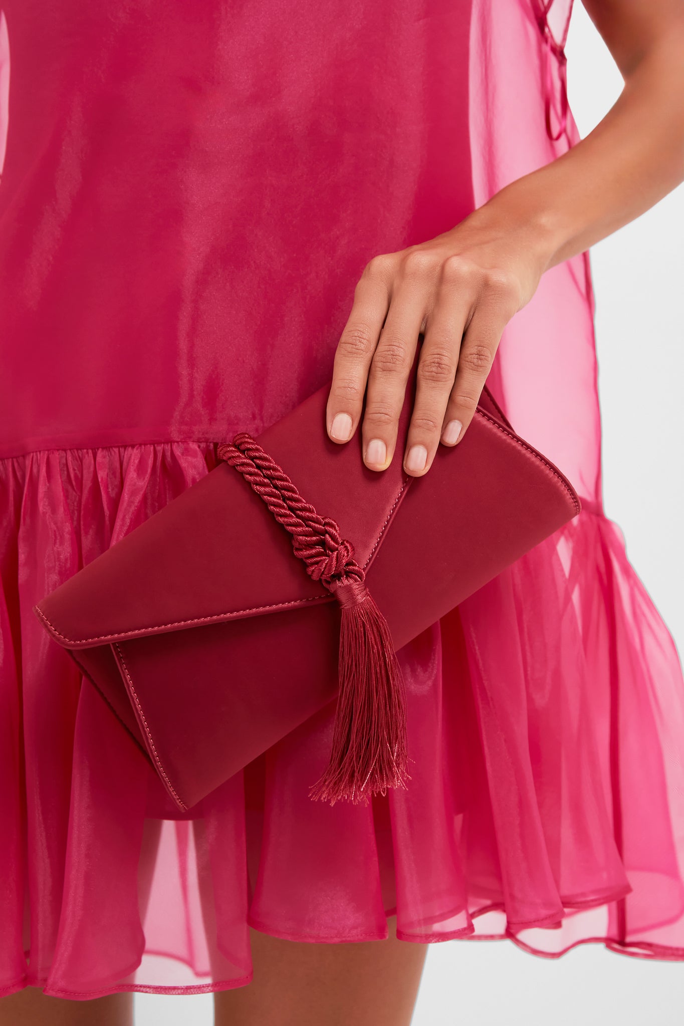 Woman wearing a sheer dress holds the classic Cranberry Satin Gabrielle Clutch, featuring an effortless envelope design and fun braided tassel, perfect for high-end occasions.