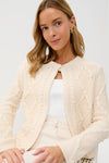 A blonde woman wears a Hyacinth House winter white cable knit cardigan with pearl detailing, paired with a white top and gold hoop earrings, seated against a plain white background.