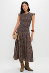 A woman stands against a white background wearing THE GREAT.’s sleeveless tapestry print midi dress with a smocked bodice, tan boots, a beige clutch, and a brown bangle.