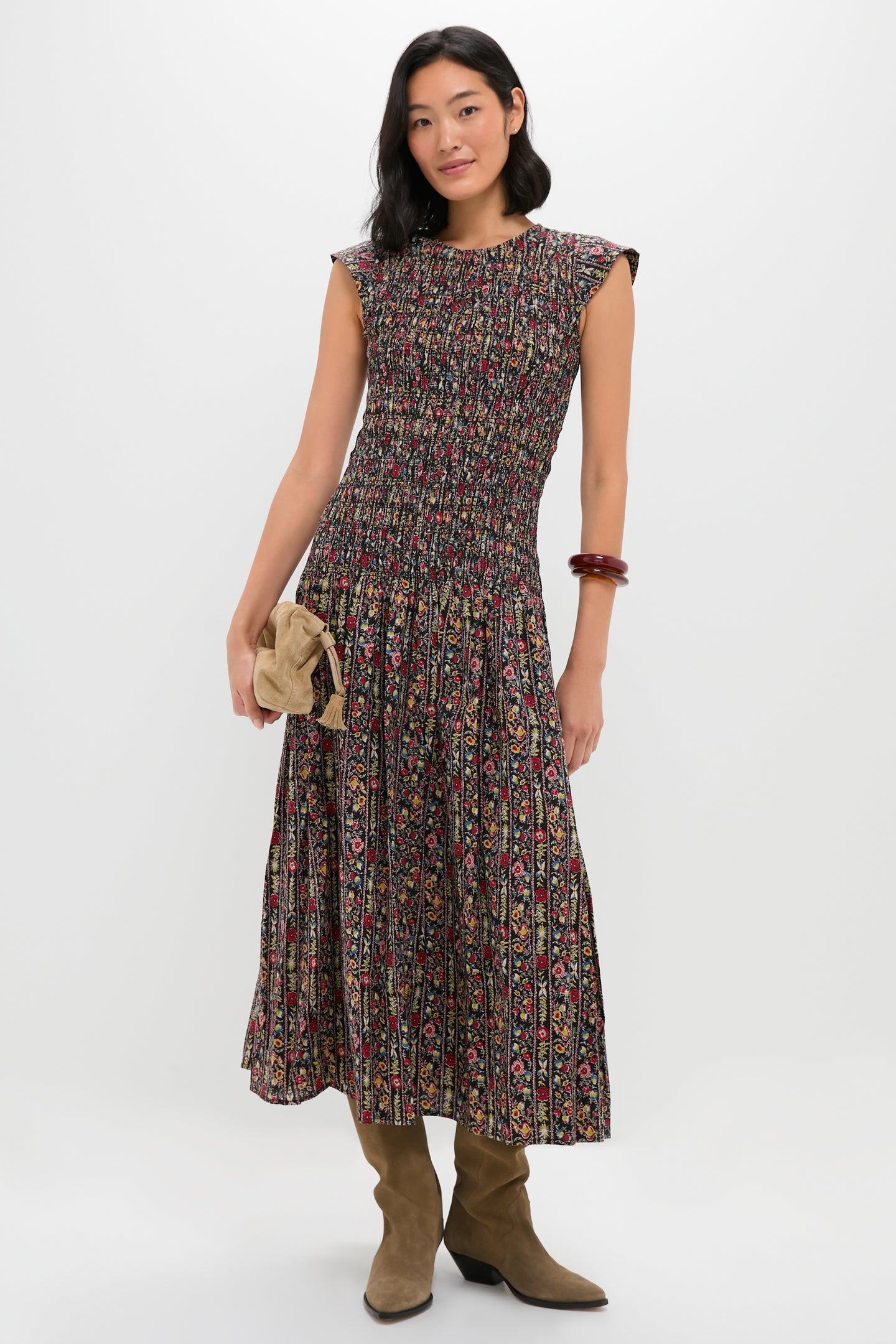 A woman stands against a white background wearing THE GREAT.’s sleeveless tapestry print midi dress with a smocked bodice, tan boots, a beige clutch, and a brown bangle.