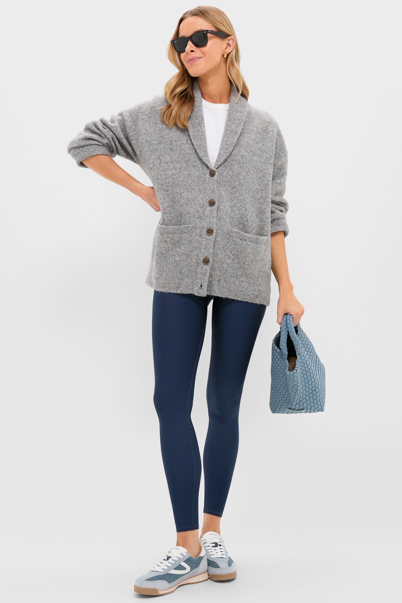 A woman in sunglasses models Tnuck Sport’s ultra-soft grey sweater, white shirt, navy leggings, and blue sneakers, holding a blue textured bag against a plain background.