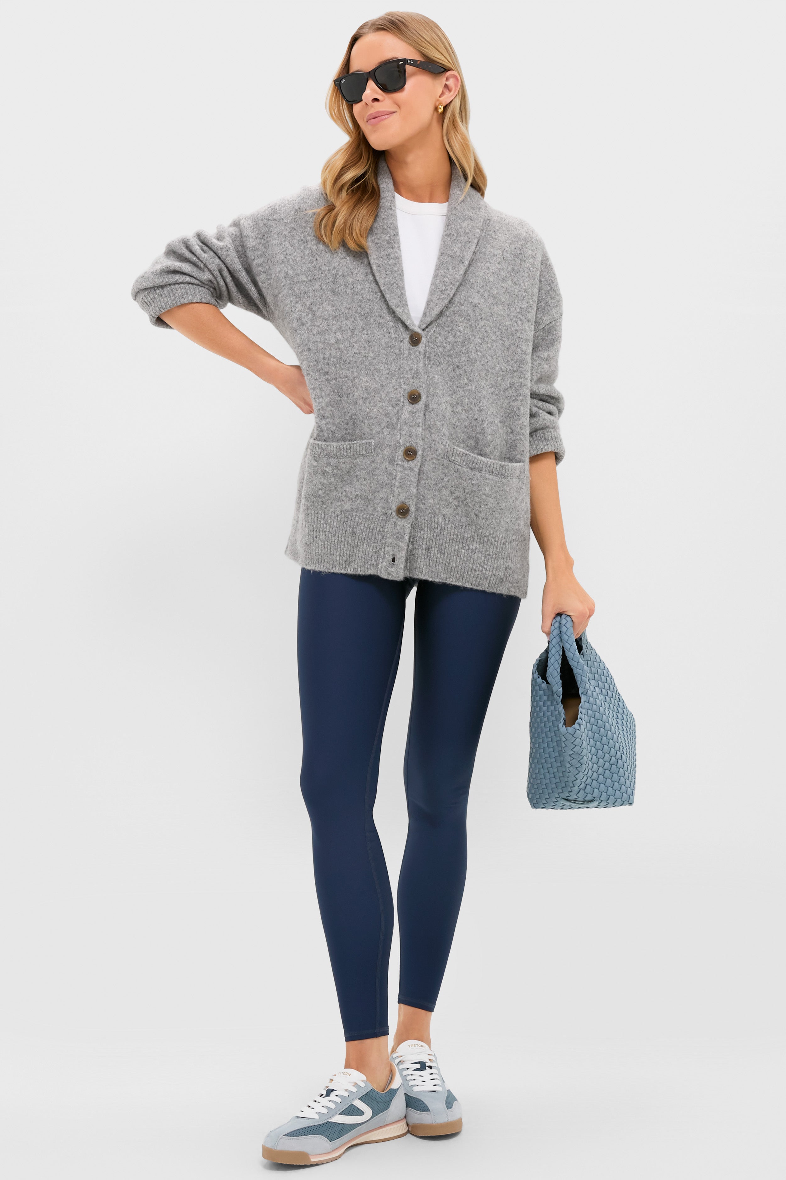 A woman in sunglasses models Tnuck Sport’s ultra-soft grey sweater, white shirt, navy leggings, and blue sneakers, holding a blue textured bag against a plain background.