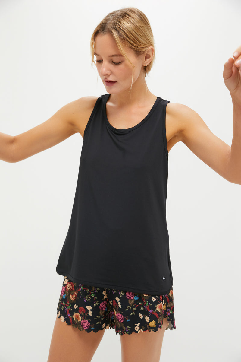 A woman stands with arms slightly raised, wearing a Tnuck Sport racerback tank and black quick-dry shorts featuring a colorful floral pattern, set against a plain light background.