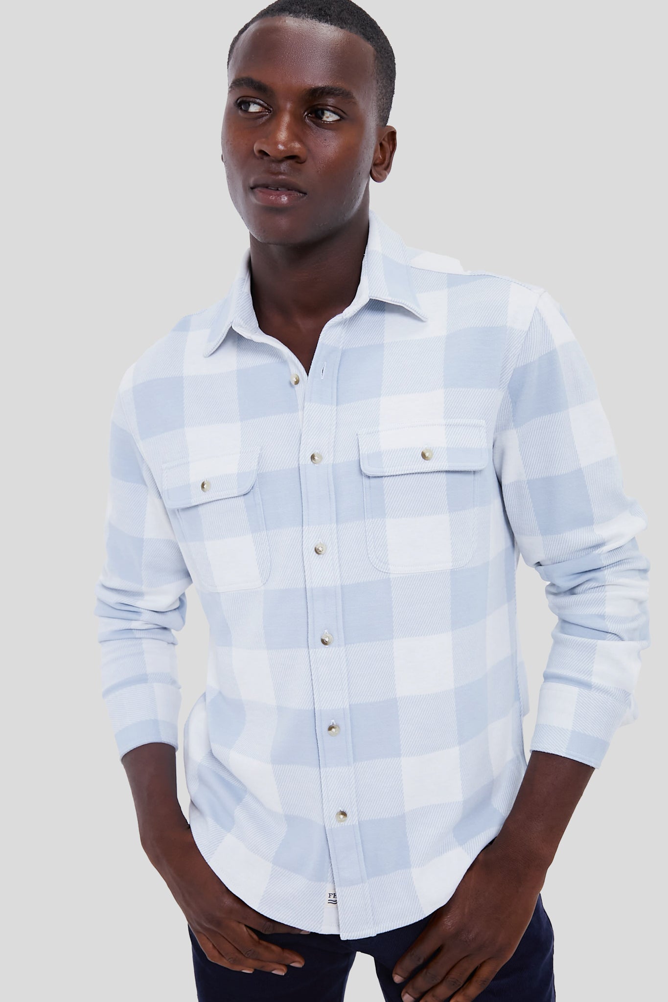 A man wears a Fair Harbor light blue plaid flannel shirt with four-way stretch, front chest pockets, and a sustainable design made from recycled plastic bottles.