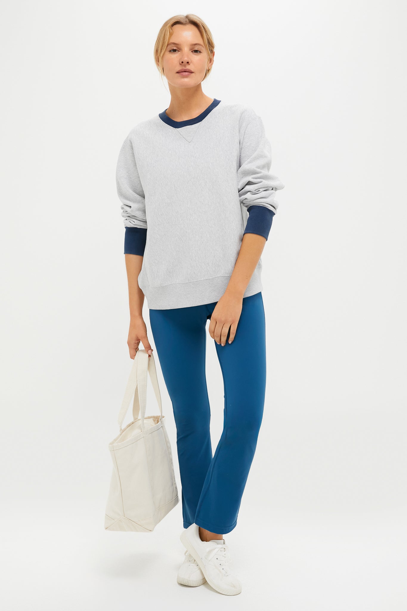 A person stands against a white background wearing a Tnuck Sport vintage-inspired crewneck sweatshirt, blue pants, white sneakers, and holding a white tote bag.