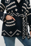 Someone wears THE GREAT. navy blue and white patterned alpaca cardigan with a belt, one hand in the pocket, styled with blue jeans.
