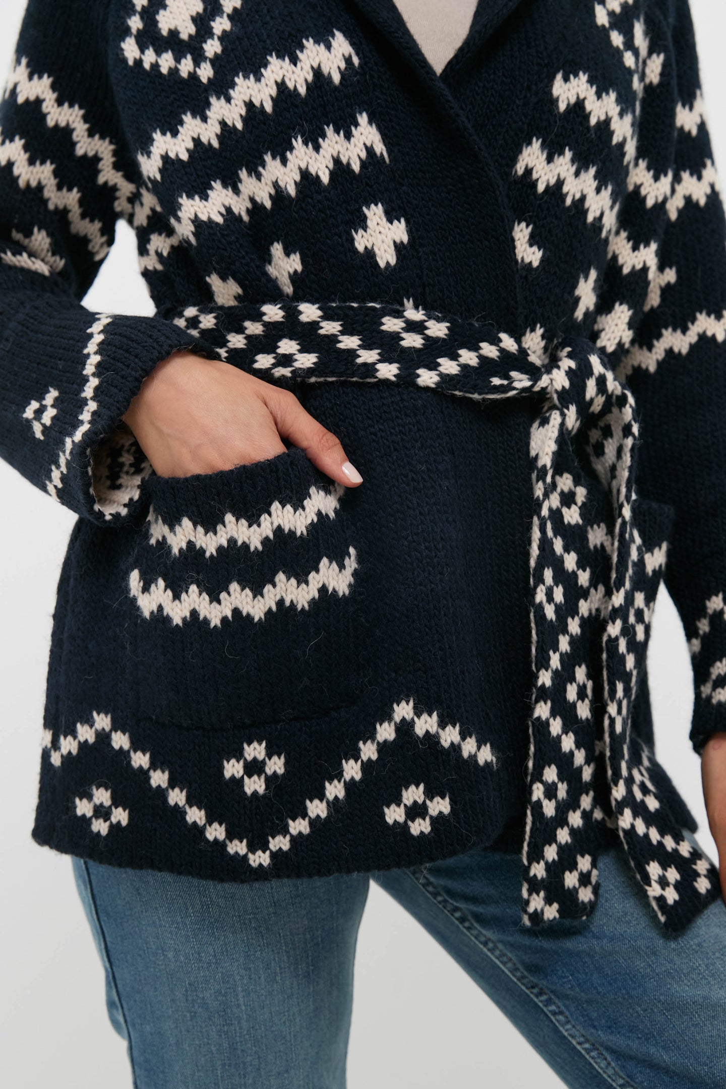 Someone wears THE GREAT. navy blue and white patterned alpaca cardigan with a belt, one hand in the pocket, styled with blue jeans.