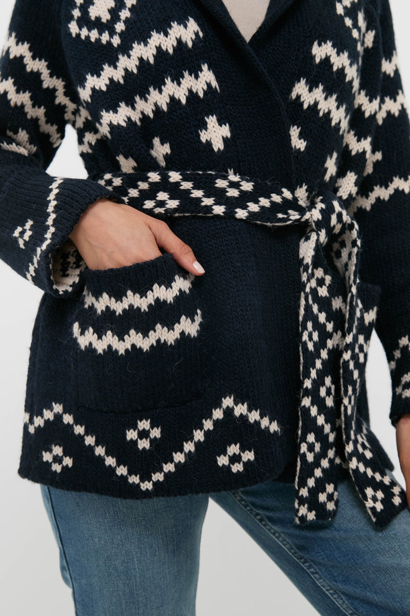 Someone wears THE GREAT. navy blue and white patterned alpaca cardigan with a belt, one hand in the pocket, styled with blue jeans.