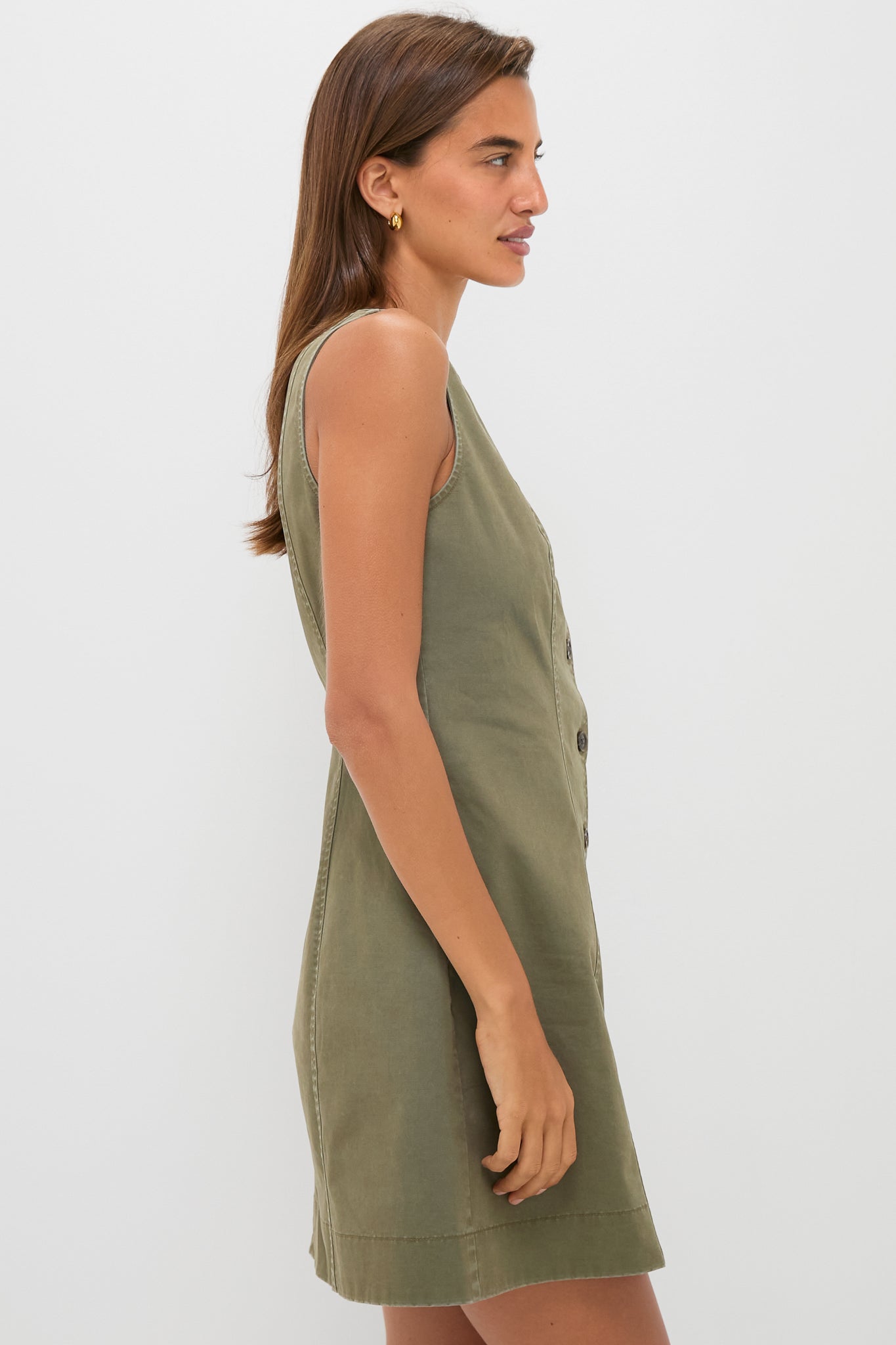 A woman stands in profile against a plain white background, wearing a Tuckernuck sleeveless olive green chino mini dress.