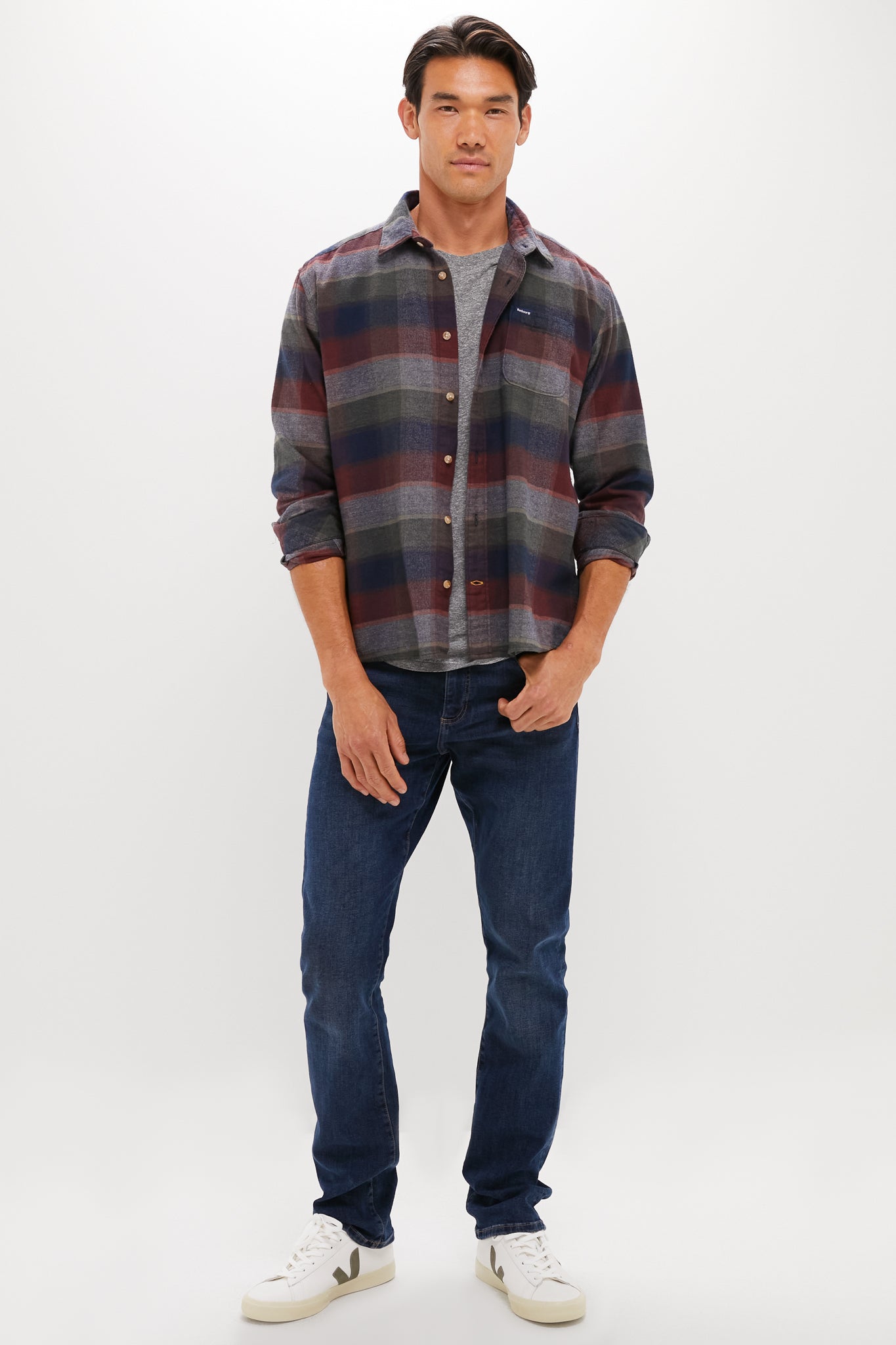 A man stands facing forward against a plain white background, wearing a Barbour relaxed long-sleeved country check shirt over a gray t-shirt, paired with dark blue jeans and white sneakers.