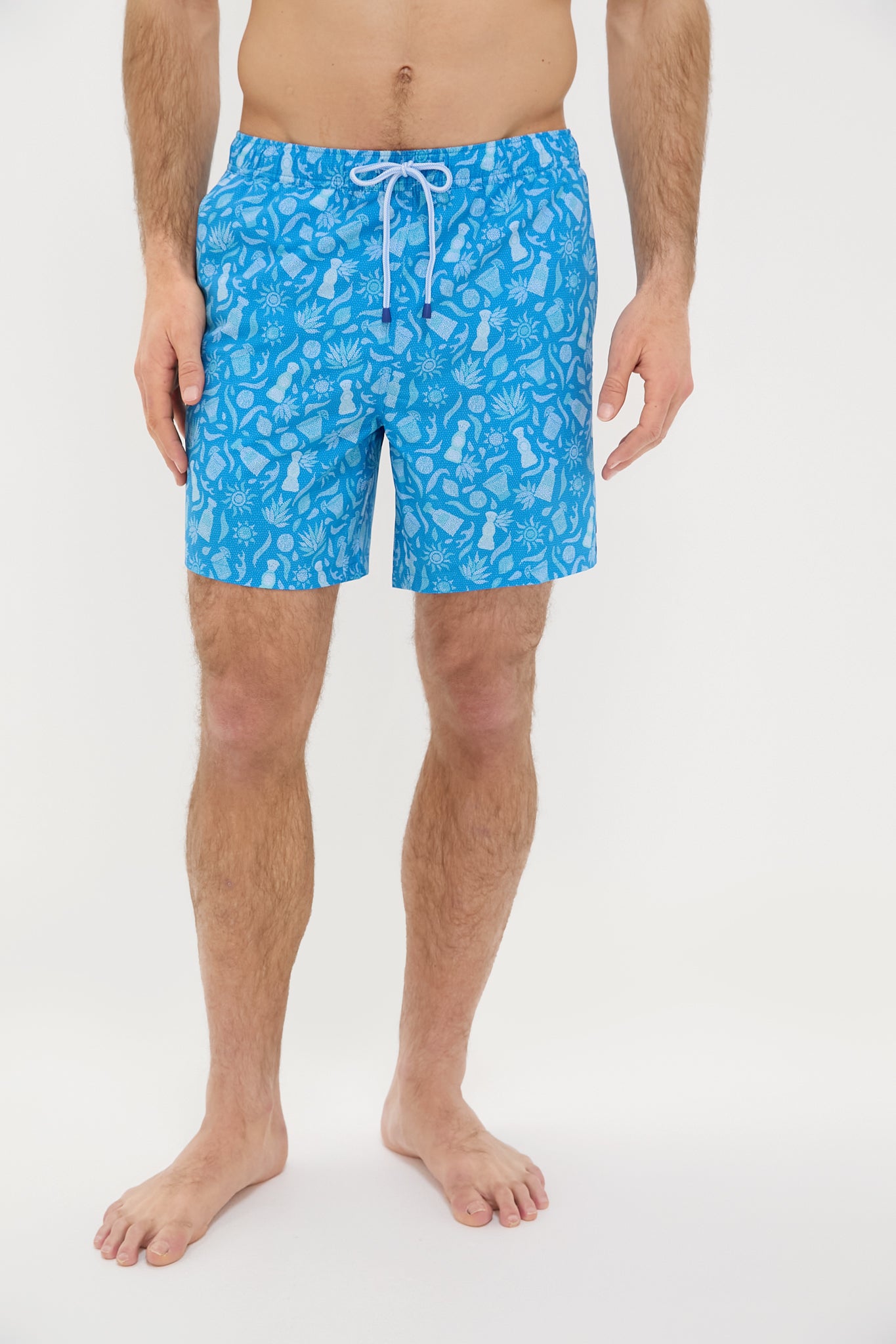 A man stands barefoot in Peter Millar Blue Macaw swim trunks featuring a white tropical leaf pattern.